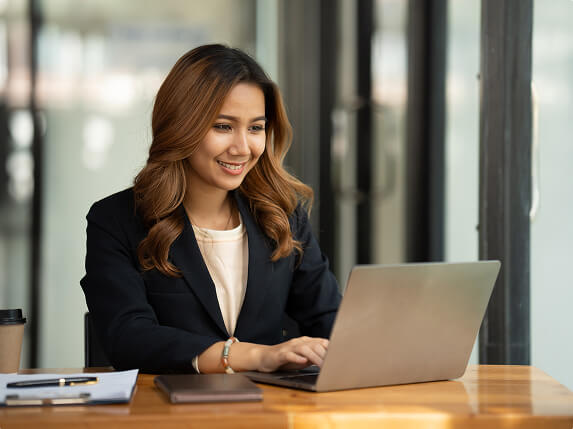 Person in blazer working on laptop at wooden desk with notebook, pen, and smartphone in modern office with large glass windows.
