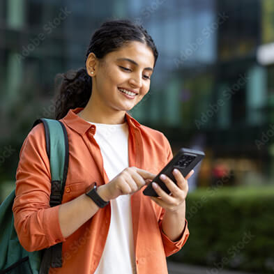 Person in orange shirt with backpack using smartphone outdoors near modern glass buildings and greenery.