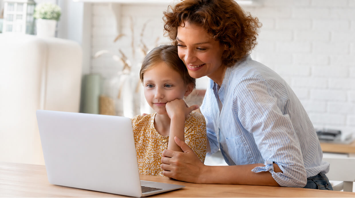 Adult and child at wooden table focused on silver laptop, striped shirt and yellow patterned shirt, white brick wall and dried plants in background.