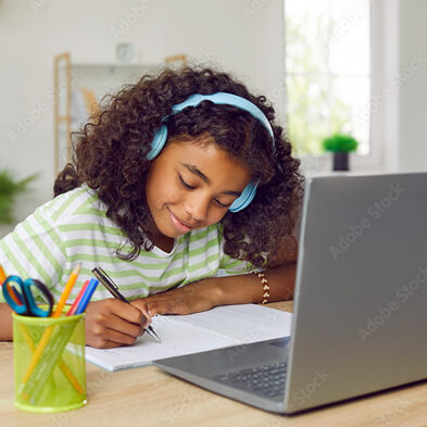 Young person with curly hair wearing headphones writing in notebook at desk with laptop and pencil holder in bright home setting.