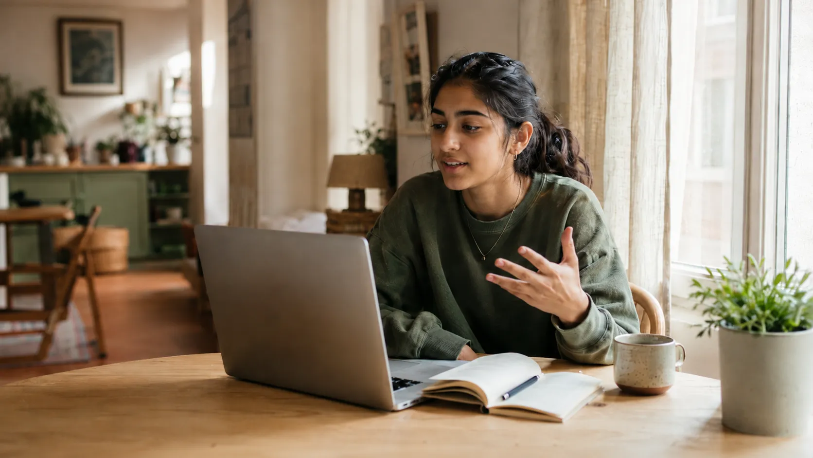 A student sitting at a desk by a window, talking during an online lesson at home.